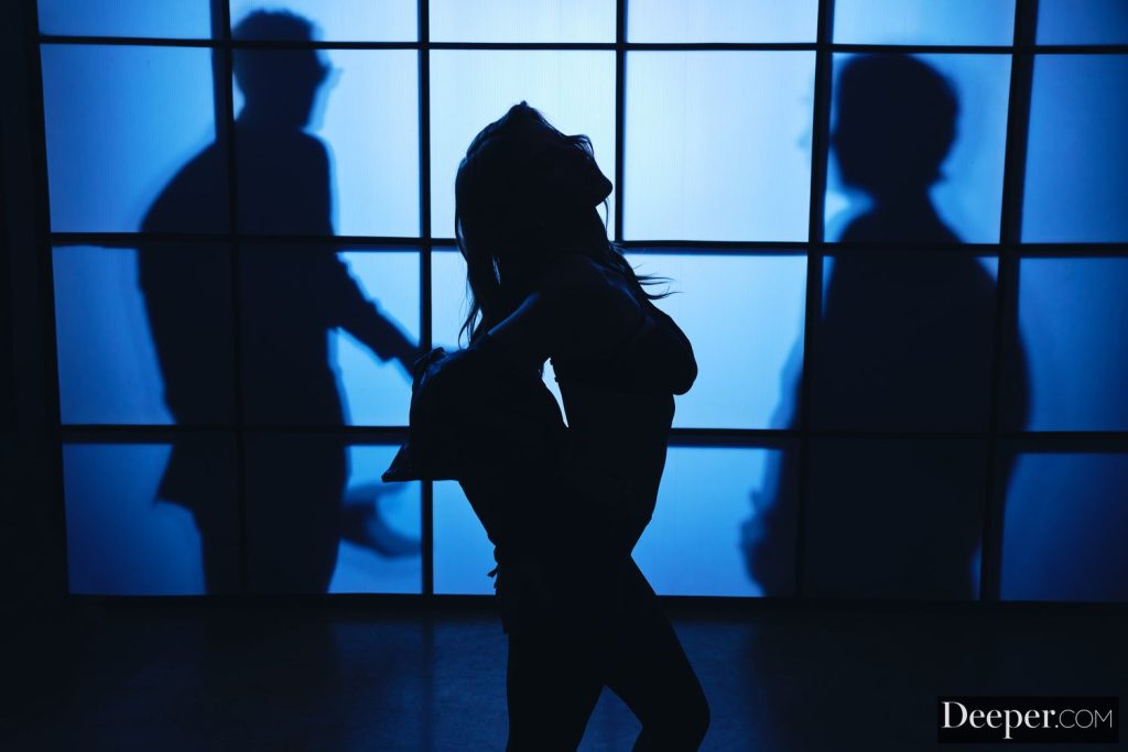 Silhouette of a woman with long hair standing in front of a blue-lit window, with the shadows of two men visible behind her.