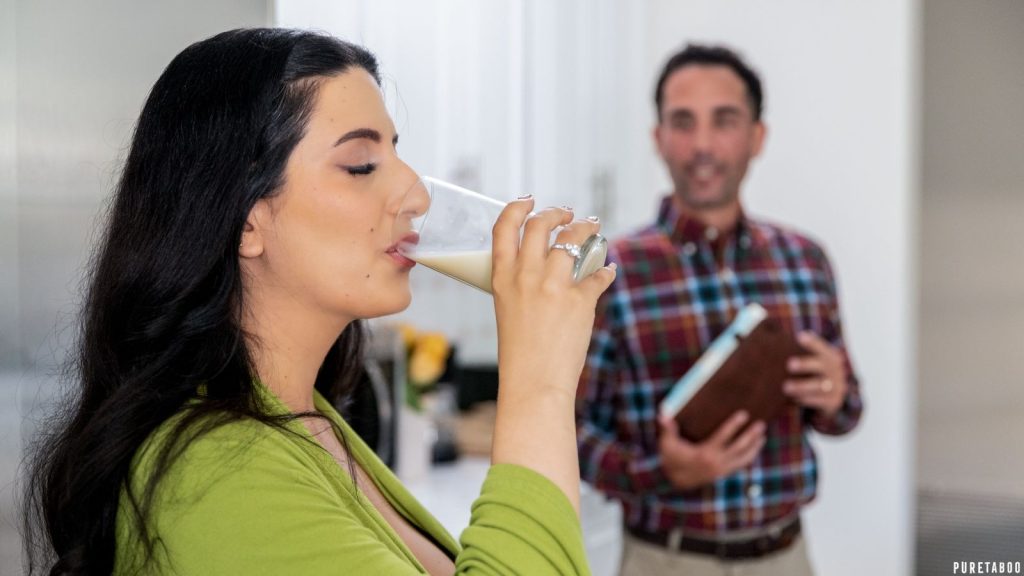 Woman with long dark hair in a green top drinking from a glass of milk in a kitchen, with a man in a plaid shirt holding a notebook in the background.