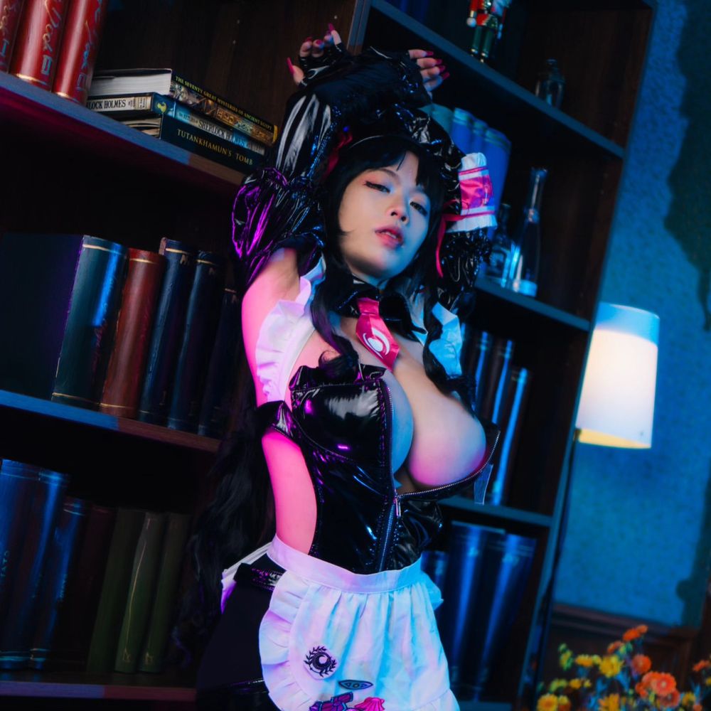 Woman with black hair in a black shiny maid-style costume with pink accents, arms raised, standing in front of bookshelves under blue and pink lighting.