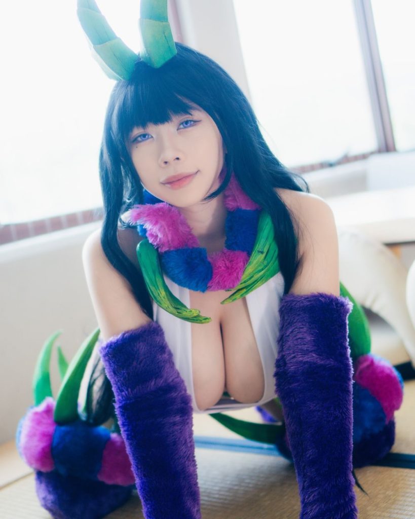 Woman with black hair and green horns kneeling on a tatami mat, wearing a white top, purple fuzzy arm and leg warmers, and a colorful fuzzy scarf, looking at the camera.