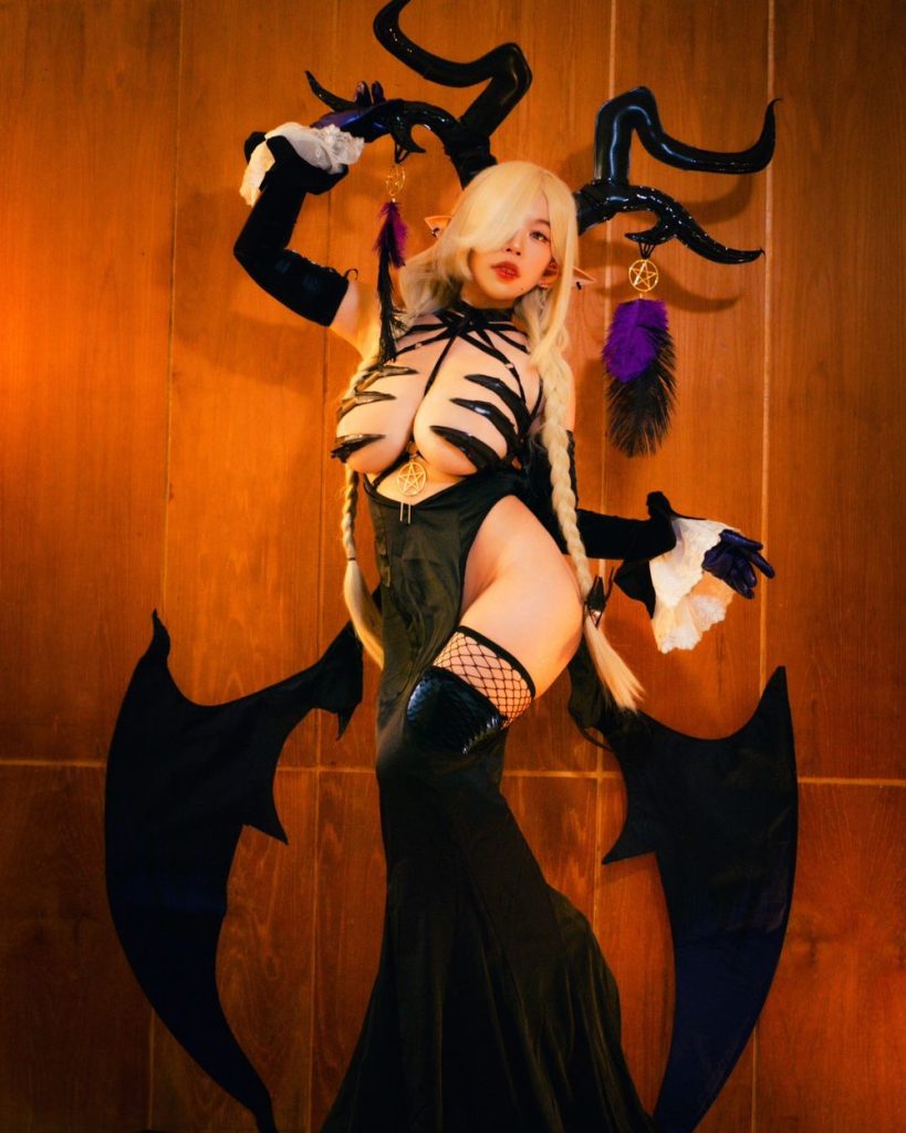 Woman with blonde hair in braids, elf ears, and large black horns, wearing a black strappy costume with wings and fishnet stockings, posing against a wooden wall with one leg raised.