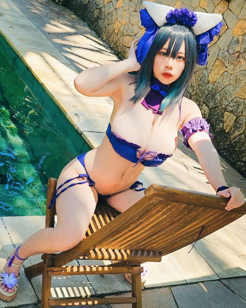 Woman with blue-tinted hair in a blue bikini and large white-and-blue headpiece, leaning on a wooden lounge chair by a pool, one hand in her hair, looking at the camera.
