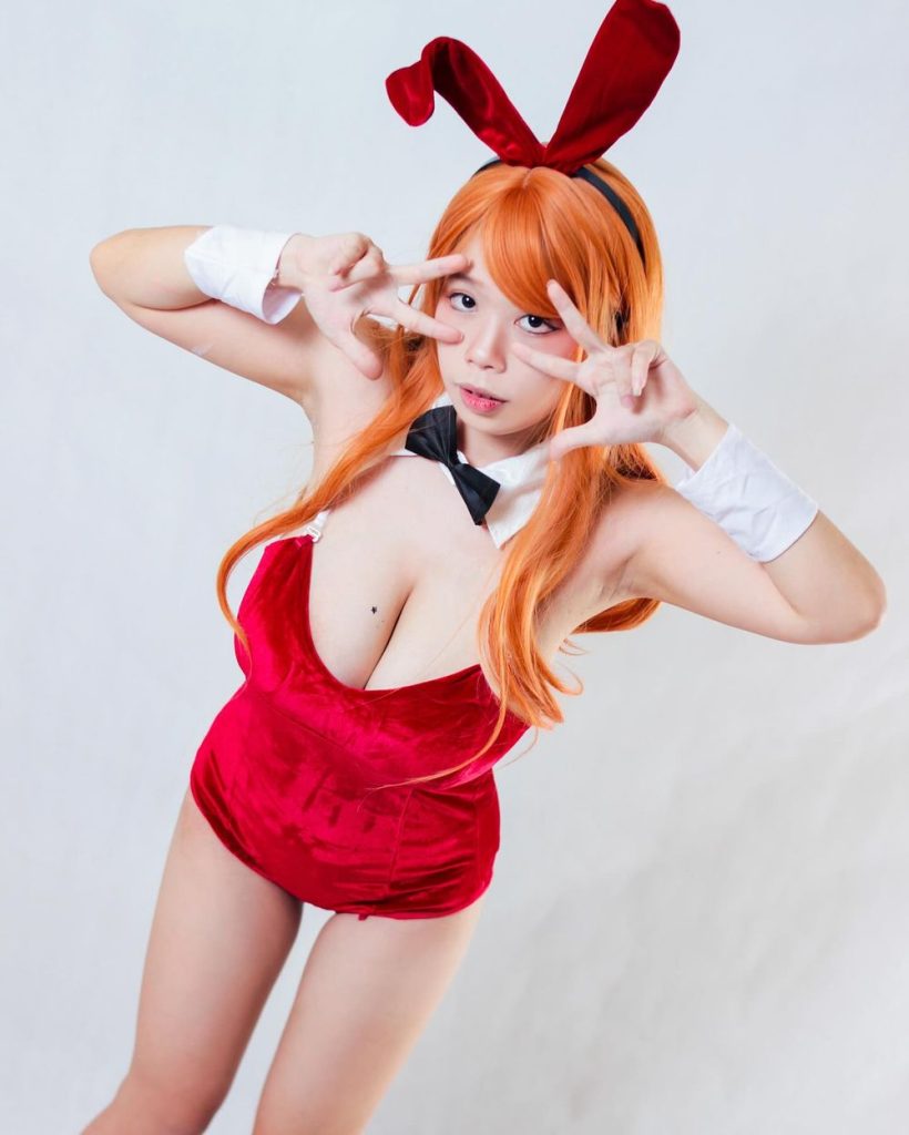 Woman with orange hair and red bunny ears wearing a red velvet leotard with white collar and black bow tie, making peace signs with both hands near her face against a plain background.