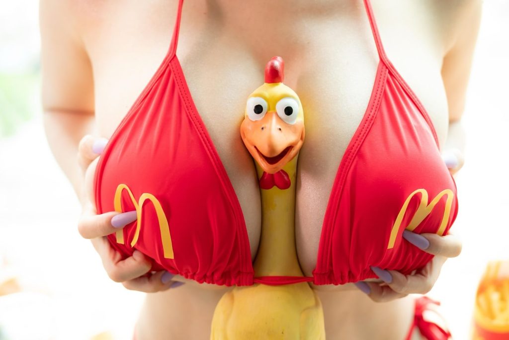 Close-up of a woman in a red bikini top with yellow McDonald’s “M” logos, holding a rubber chicken between her breasts.