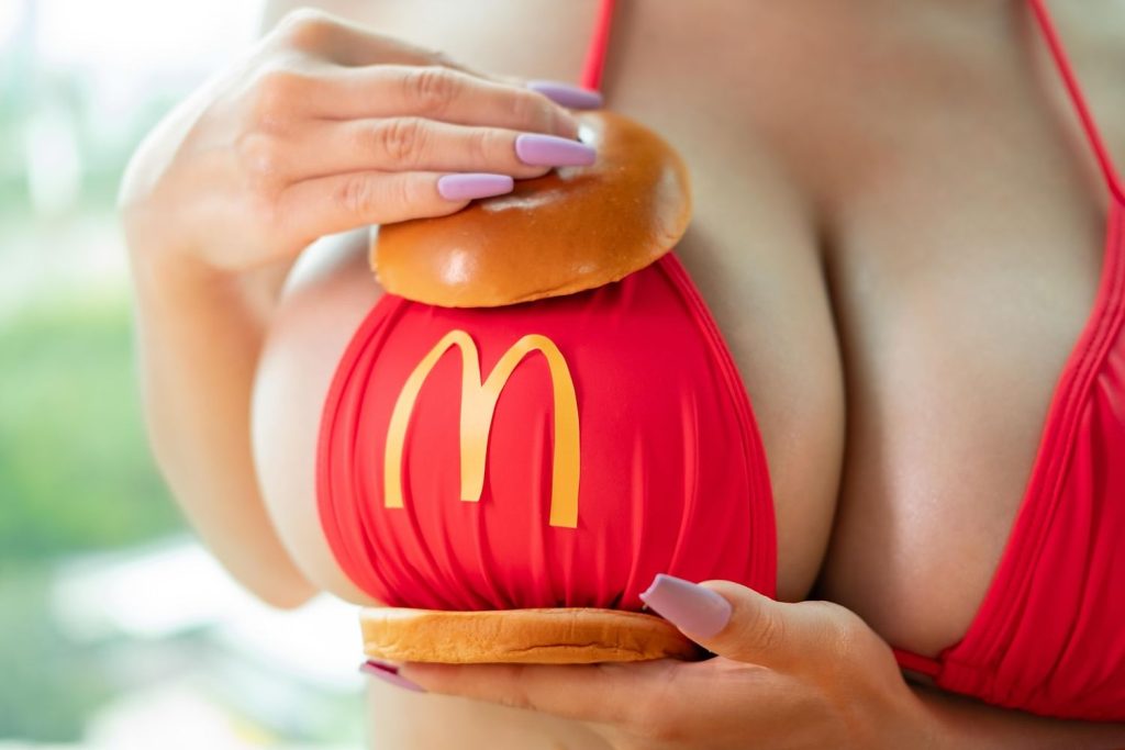 Close-up of a woman in a red bikini top with yellow McDonald’s “M” logo, holding a hamburger bun against one breast.